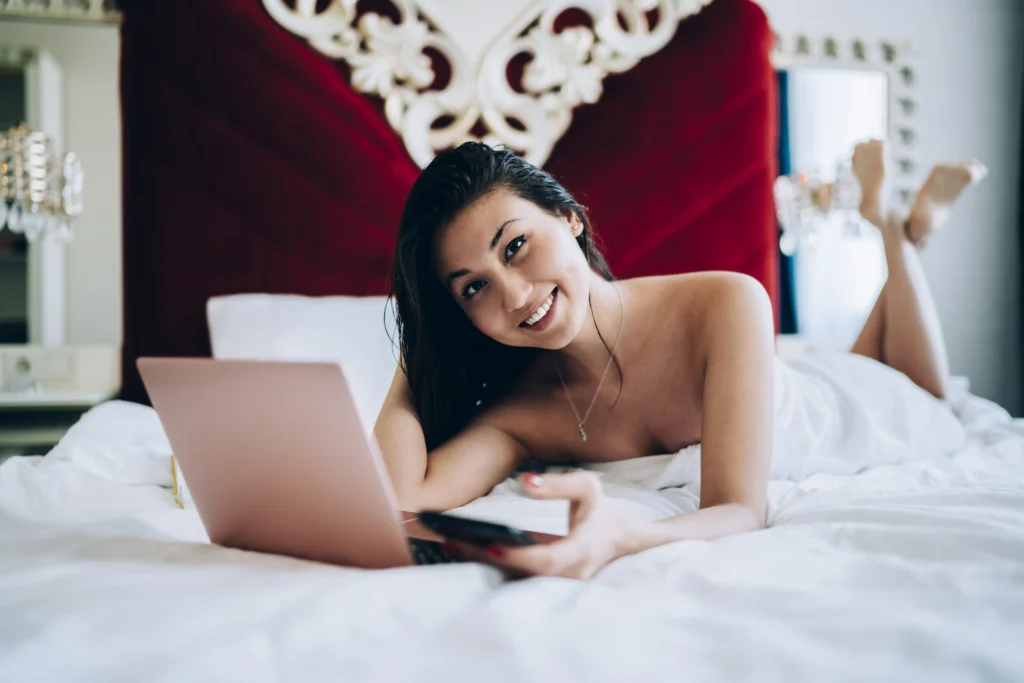 Chica sonriente recostada en una cama mientras utiliza una laptop para chatear en línea.