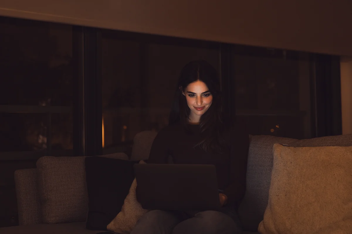 Chica trabajando de noche desde el sofá con su portátil en un empleo de chatter en OnlyFans